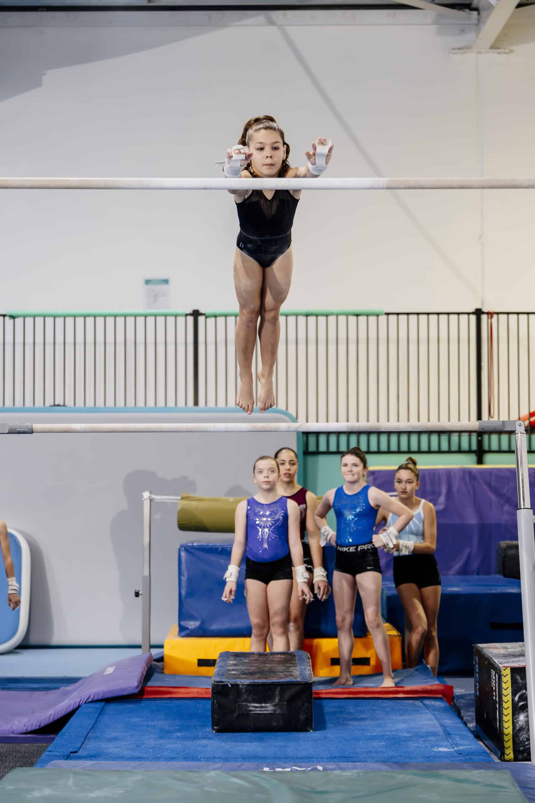 Junior gymnast jumping to high bar during training, teammates watching from below